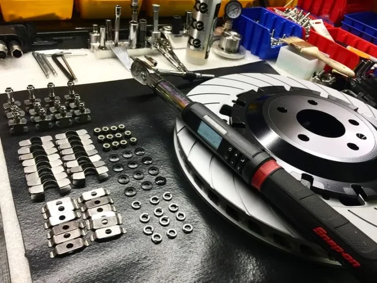 A workbench with organized metal fasteners and small parts next to a torque wrench placed on top of a slotted brake rotor, with tools and bins visible in the background.