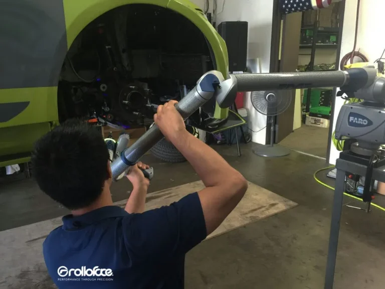 A Rolloface technician holding a portable measurement arm (FARO) up to a vehicle’s wheel well inside a garage setting, with the vehicle partially disassembled and tools visible in the background.